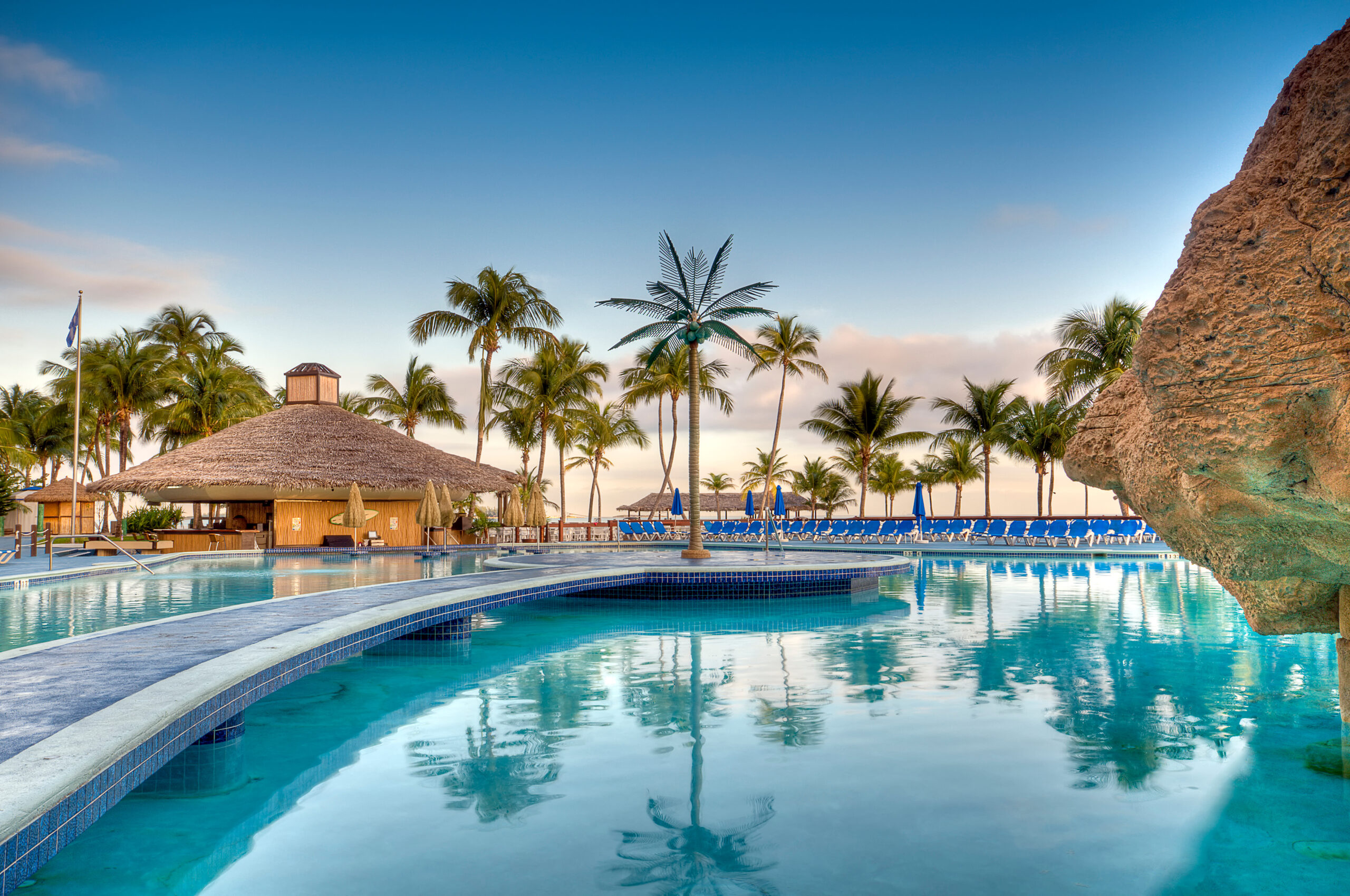 Luxury tropical resort pool with palm trees, thatched-roof pool bar, lounge chairs, and calm turquoise water under a clear blue sky.